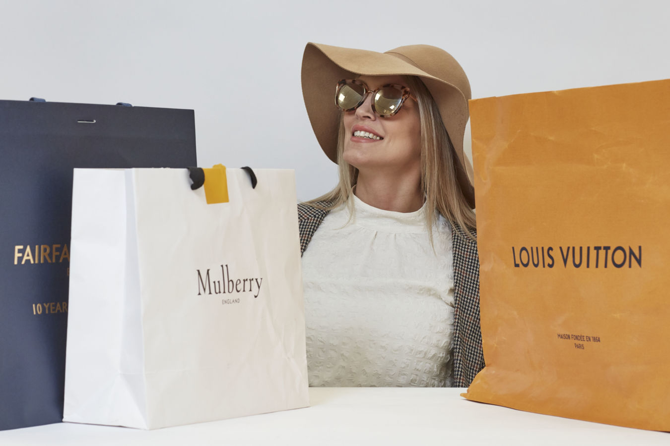 Woman smiling, wearing a tan hat and sunglasses, surrounded by luxury shopping bags labeled Fairfax, Mulberry, and Louis Vuitton on a table, in a minimal retail setting.