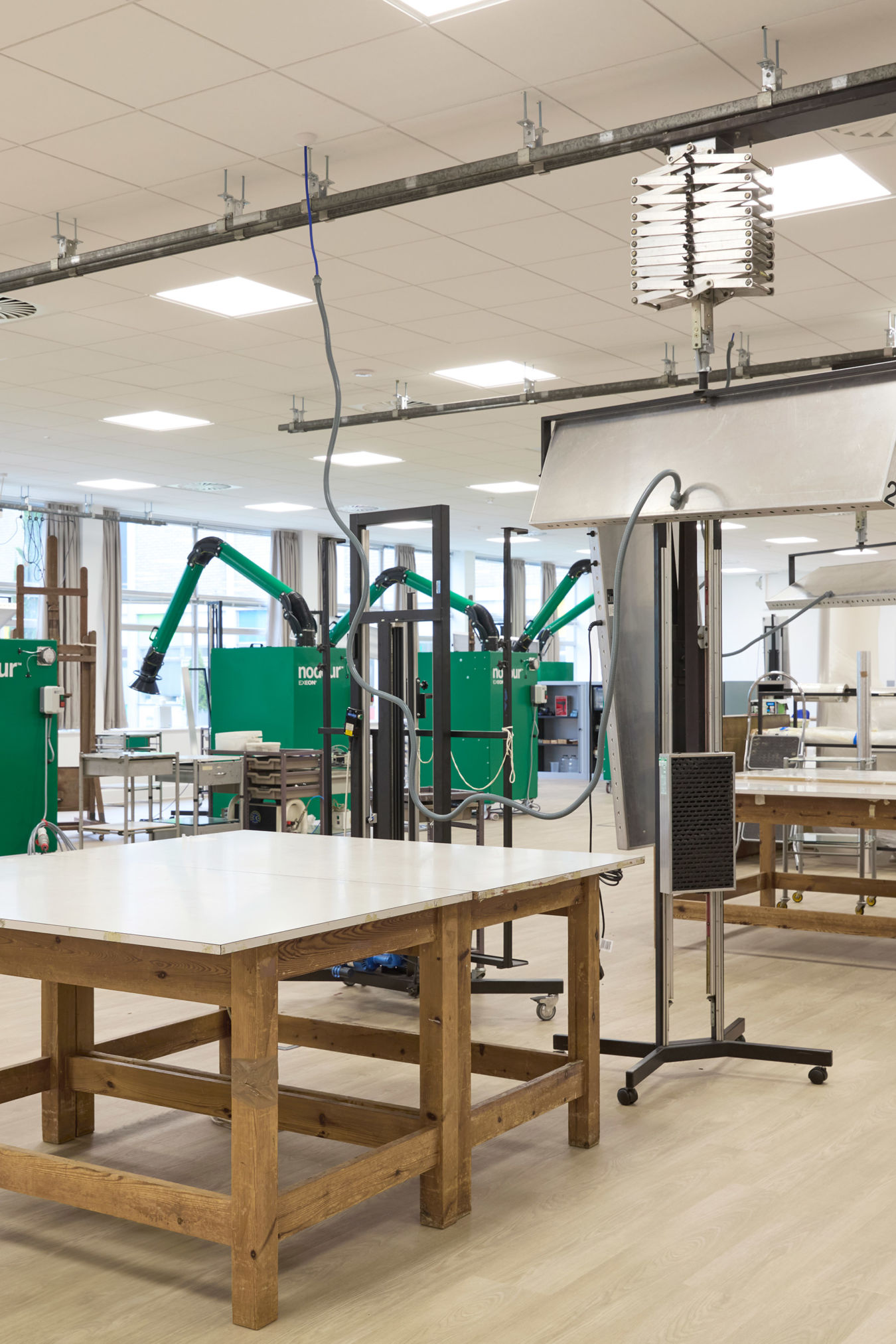 Spacious workshop with wooden tables features green ventilation units labeled "nodour" and extendable hoses, set in a bright room with white ceiling panels and large windows.