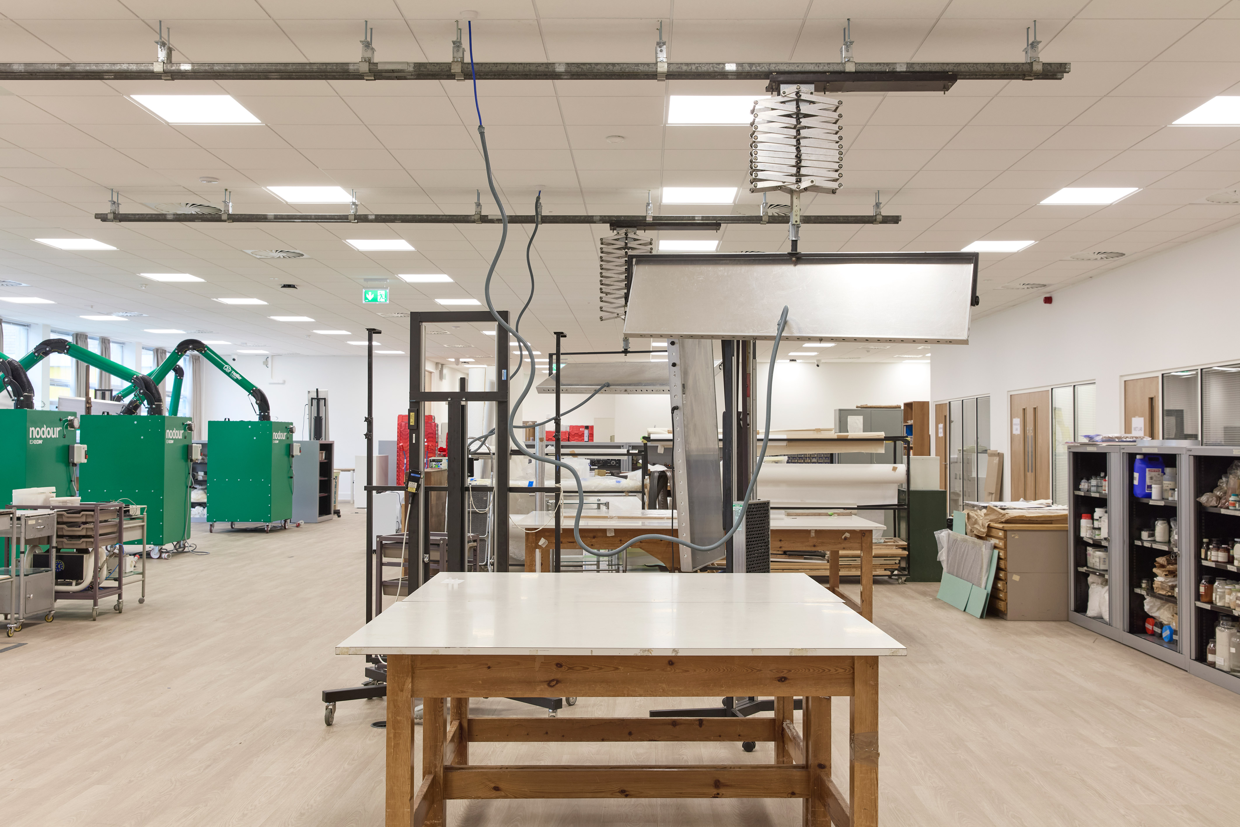 A spacious workshop with a wooden table under suspended equipment; "nodour" machines with articulated arms line one side, shelves with supplies on the other, and bright ceiling lights above.