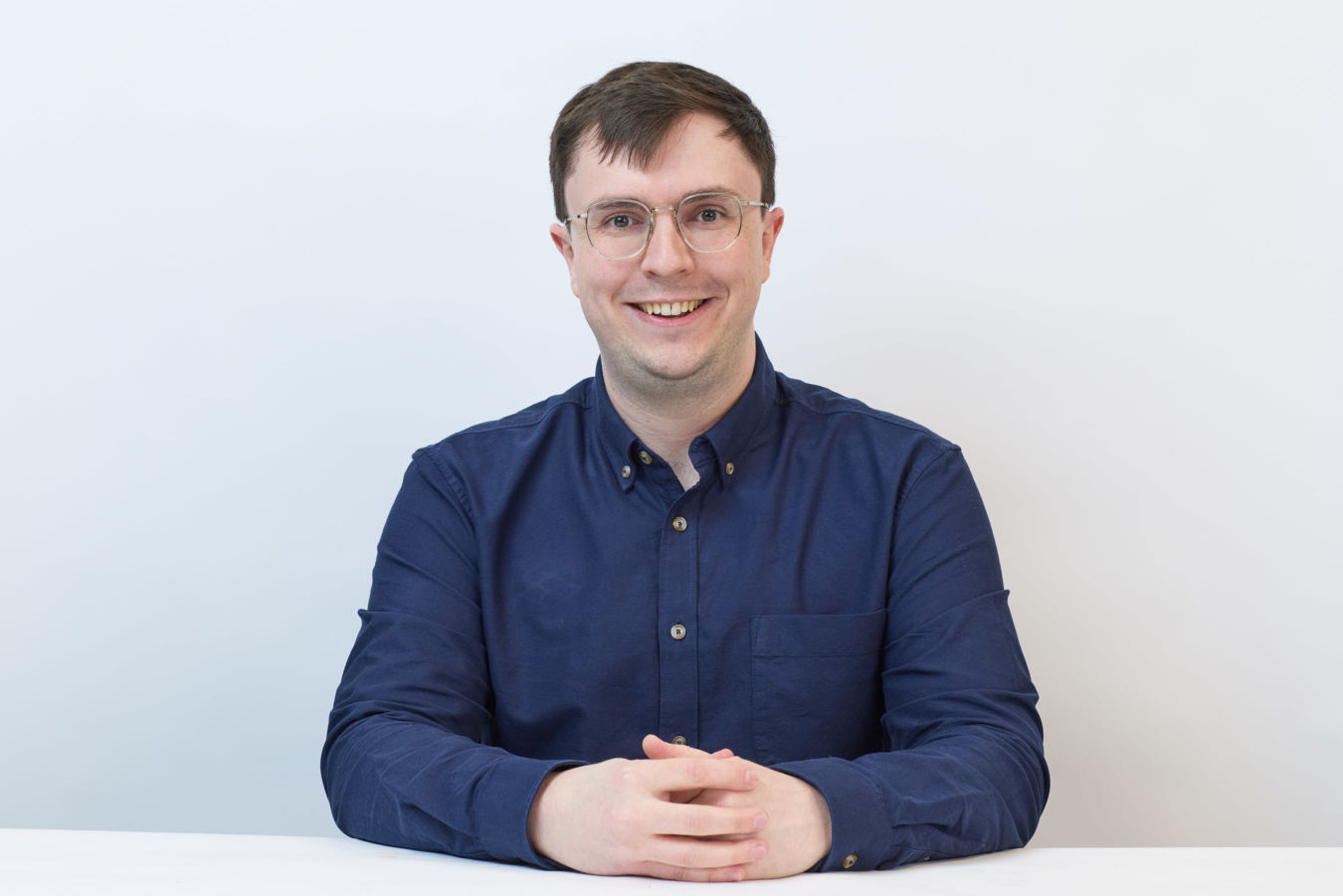 A smiling person wearing glasses and a blue button-up shirt sits with hands clasped on a white table against a plain white background.