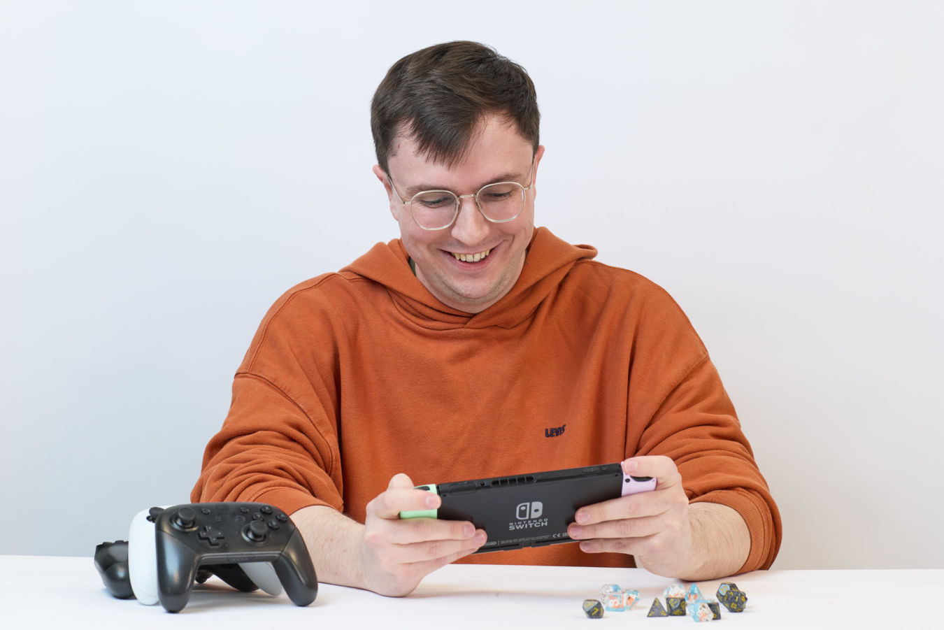 Person smiling while playing Nintendo Switch, wearing an orange hoodie, seated at a table with colorful dice and a controller nearby in a simple, light setting.