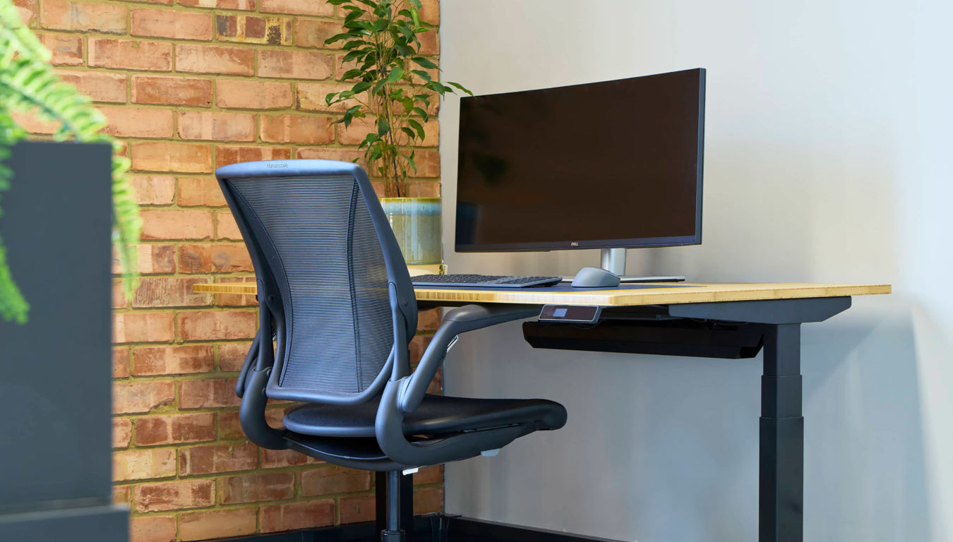 A modern workspace with an ergonomic chair and a wooden desk featuring a large monitor, keyboard, and mouse. A brick wall and plant add warmth.
