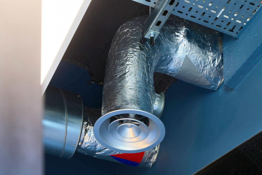 entilation duct with shiny silver insulation and a circular grille is attached to a blue ceiling. The setting is bright and industrial.