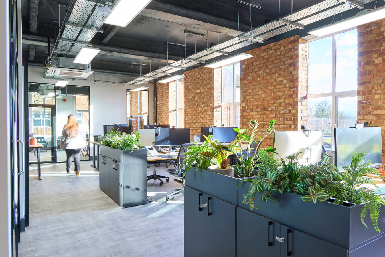 Modern office with large windows, brick walls, and sleek furniture. Green plants add freshness. A person is walking, conveying a lively atmosphere.
