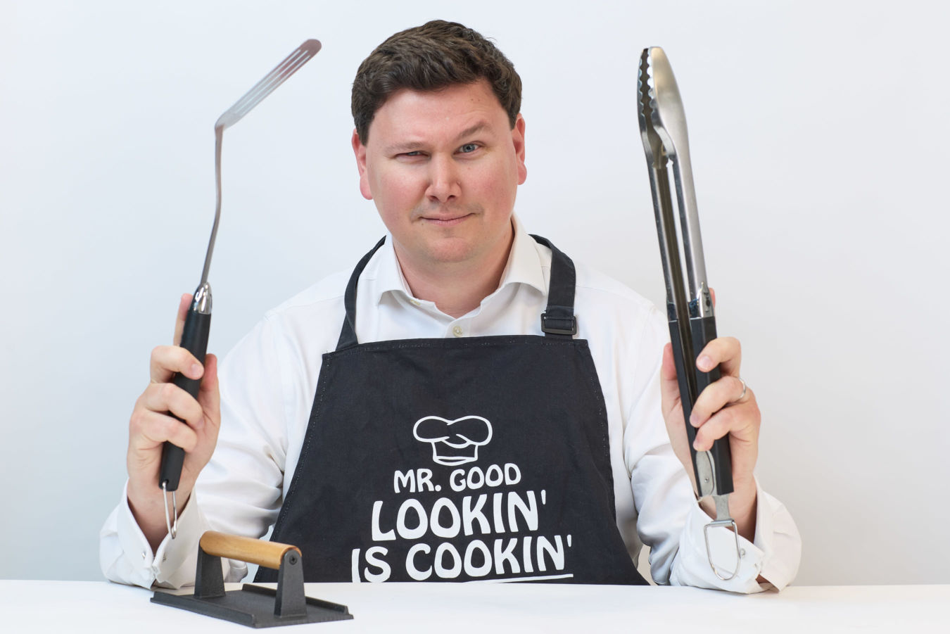 A person wearing an apron reading "MR. GOOD LOOKIN' IS COOKIN'" holds grilling tools, smiling, at a table, against a plain white background.