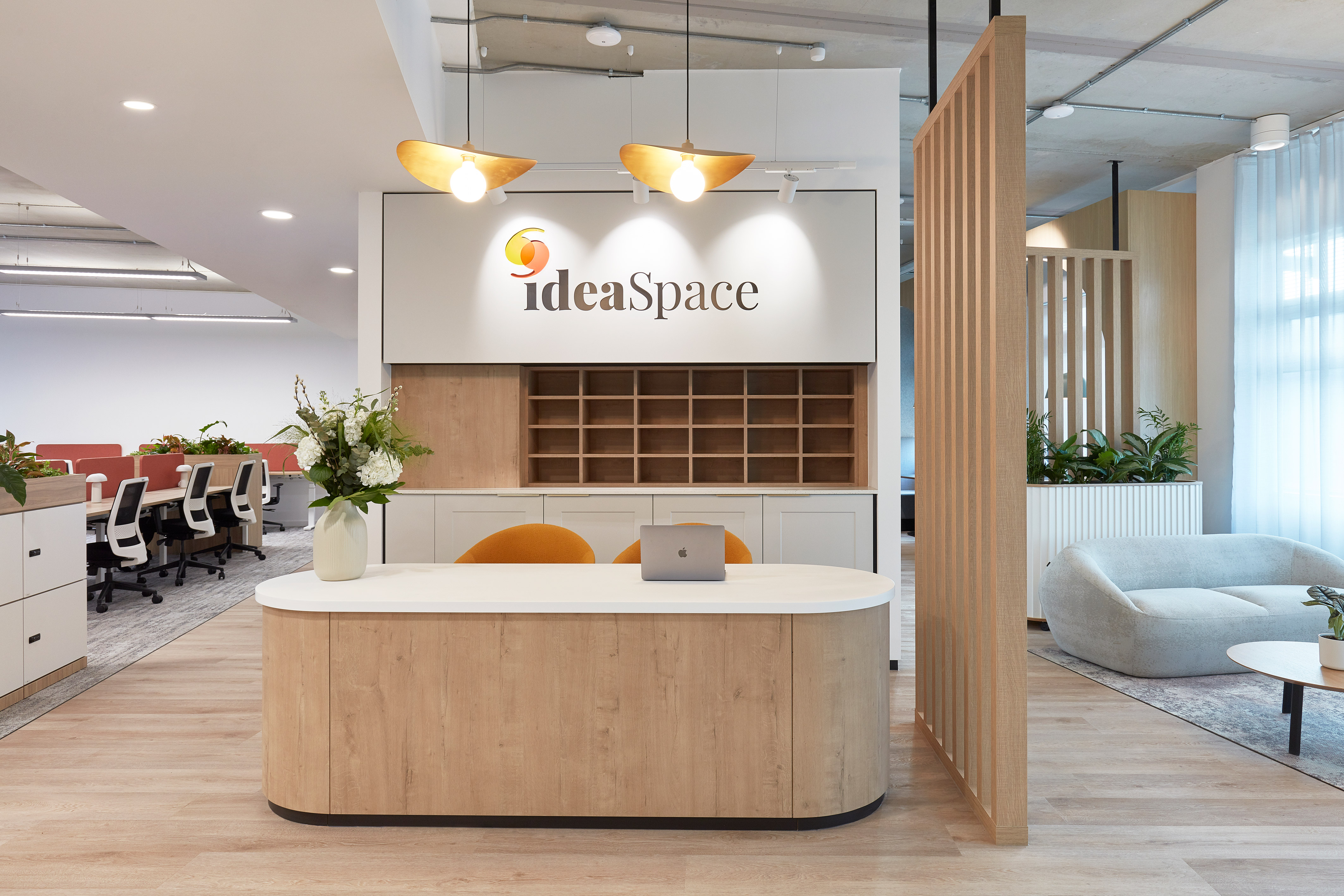 Reception desk with a laptop and flowers beneath the "ideaSpace" logo, in a modern office featuring wooden floors, partition panels, chairs, and large windows.