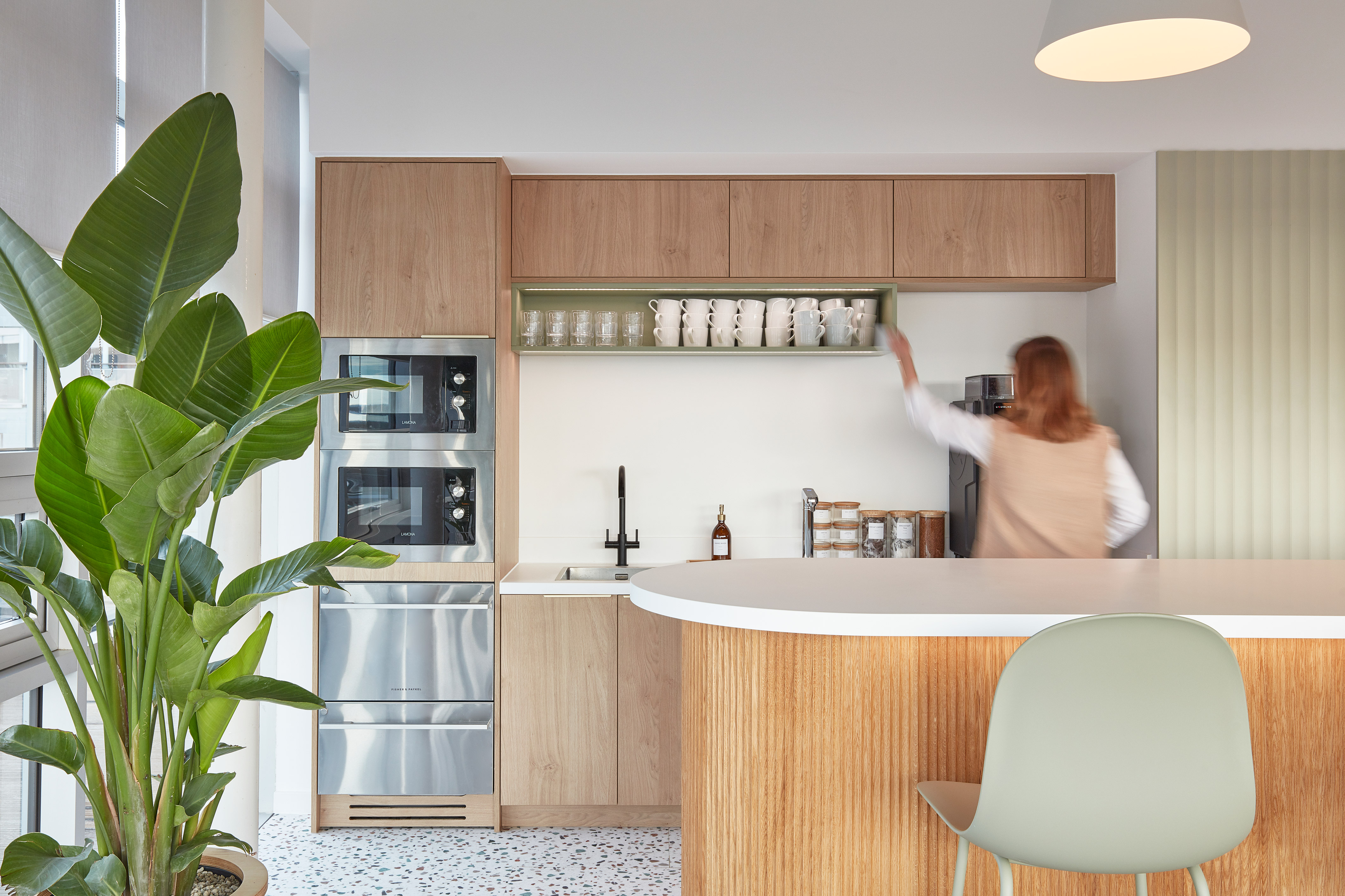 A modern kitchen with a person reaching for cups on a shelf above a sink, surrounded by wooden cabinets, a plant, and two built-in appliances.