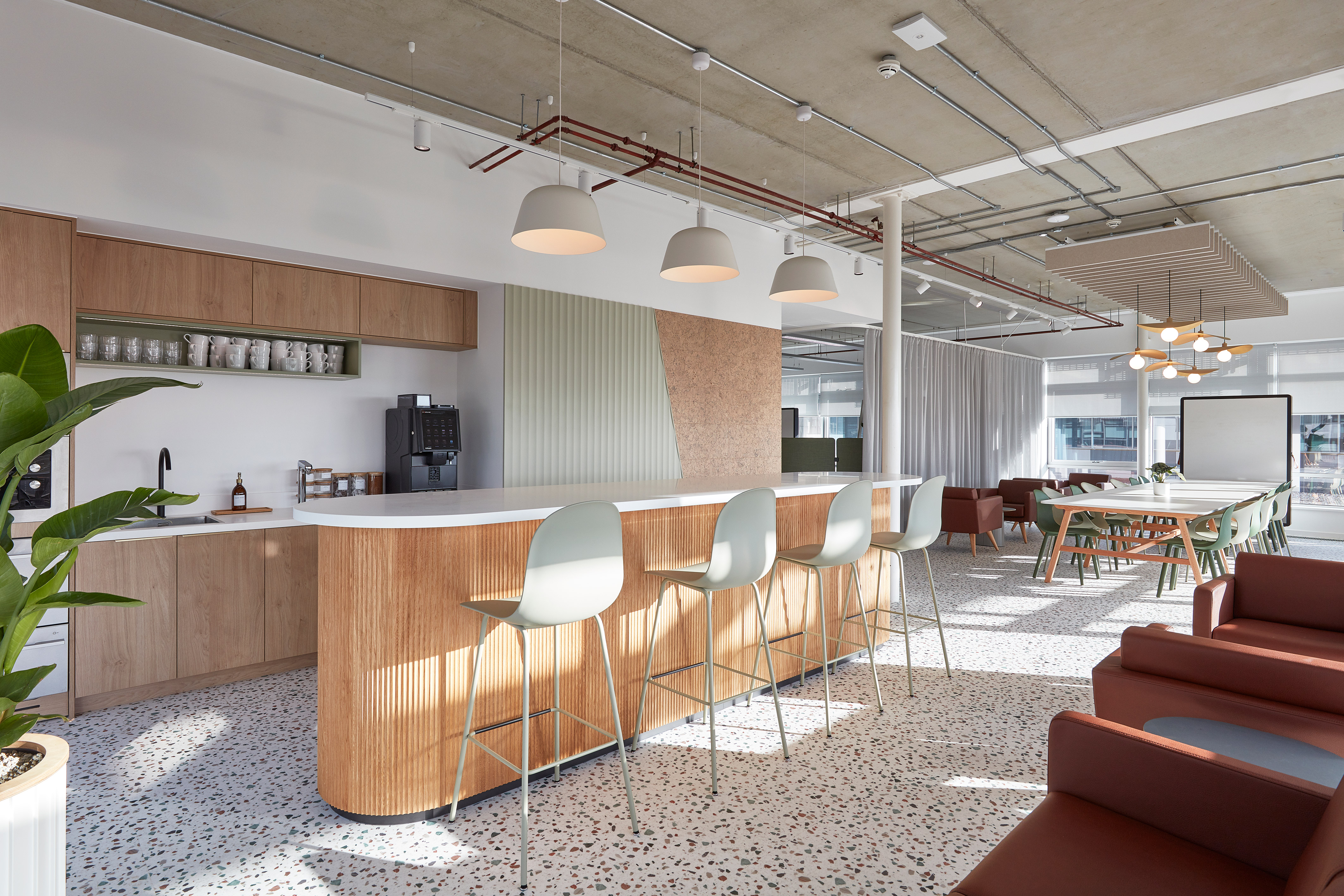 Modern office lounge with a wooden counter and light blue chairs, surrounded by brown sofas and large tables. Pendant lights hang from the ceiling, with a plant and windows in the background.