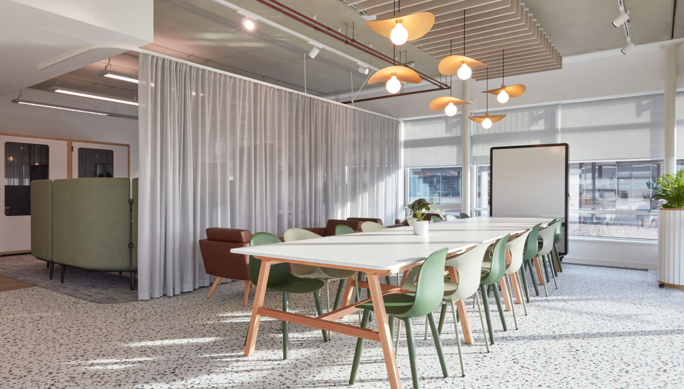 Modern office space with large white tables and green chairs. Ceiling lamps hang over tables. A curtain divides the space, creating a cozy, open atmosphere