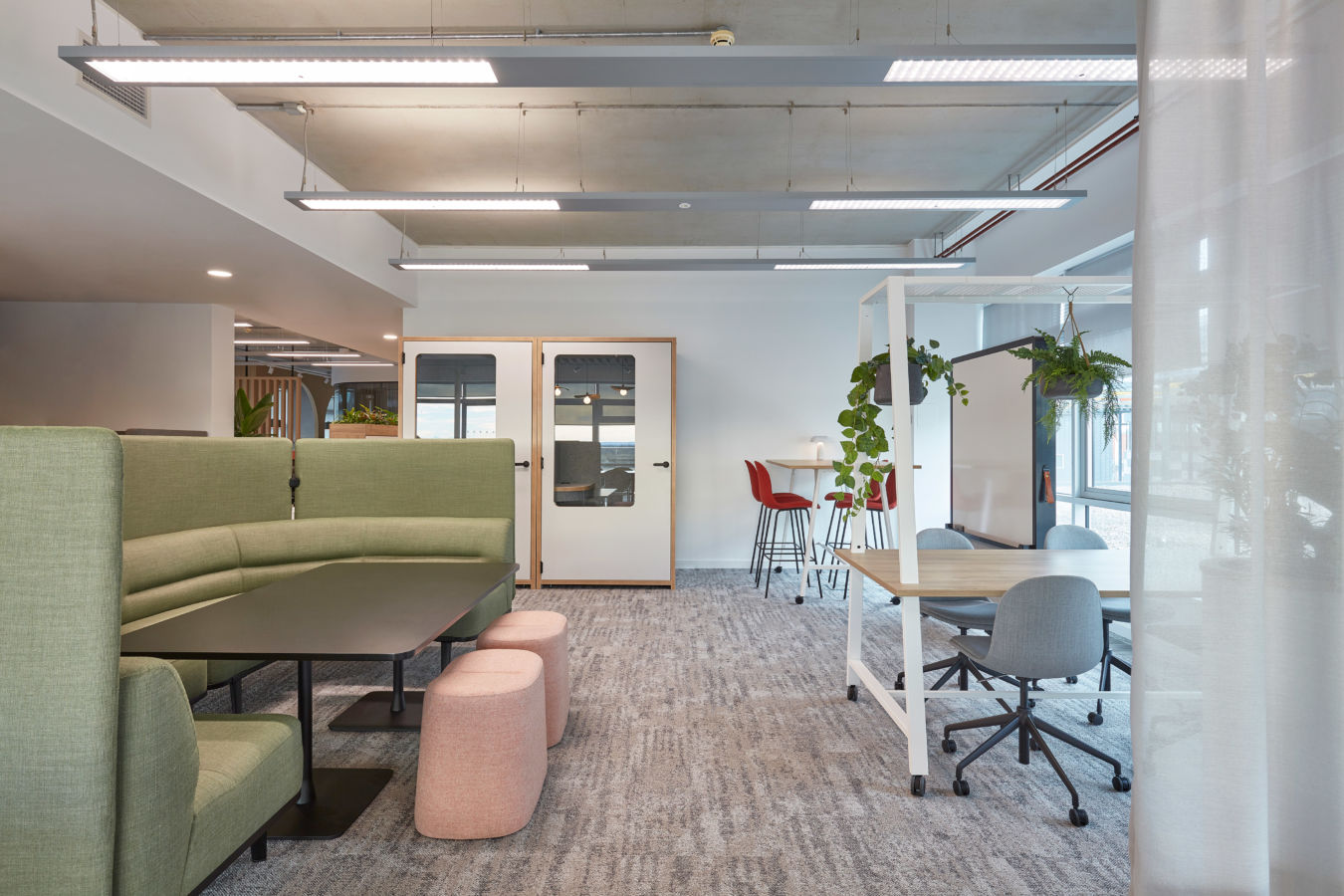 Green sectional sofa with a black table and pink stools is placed in a modern office space. Nearby, red chairs and plants adorn the bright, open environment.