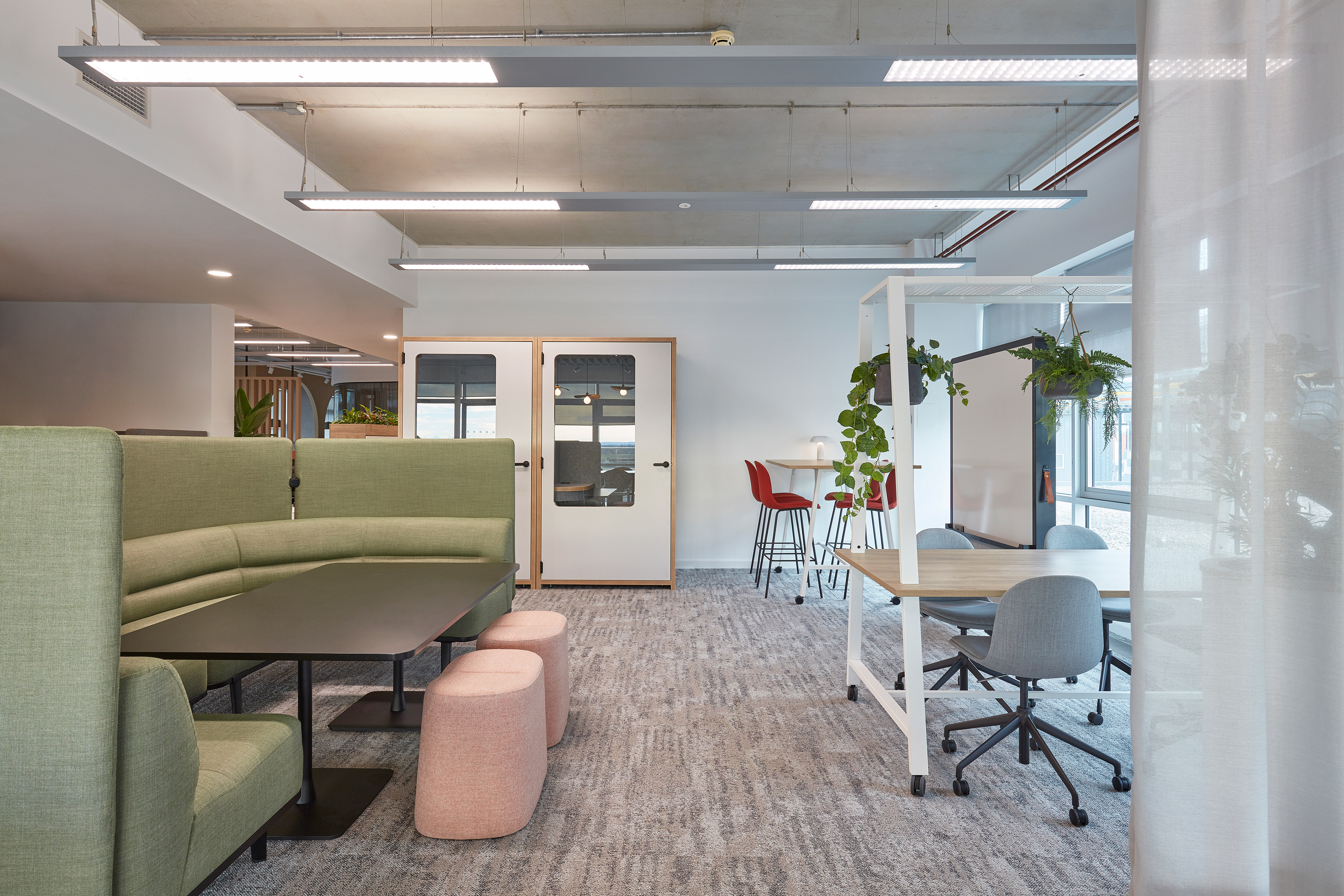 Green sectional sofa with a black table and pink stools is placed in a modern office space. Nearby, red chairs and plants adorn the bright, open environment.