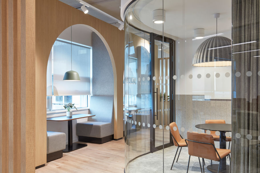 office with a cozy seating nook under an arched wooden frame, featuring gray cushions, a hanging lamp, and a small plant. Curved glass wall encloses a meeting space, adding a sleek, inviting atmosphere