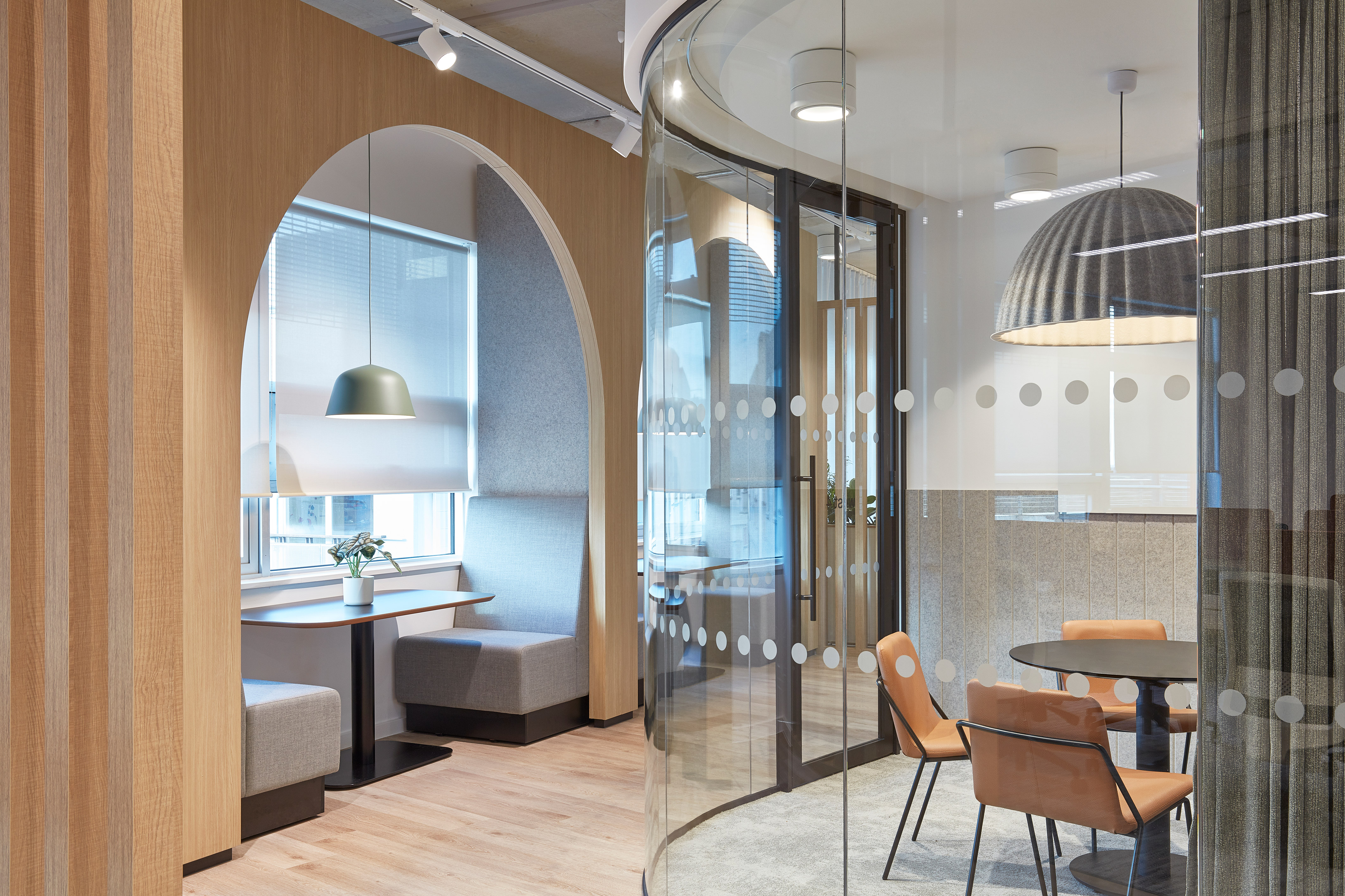 office with a cozy seating nook under an arched wooden frame, featuring gray cushions, a hanging lamp, and a small plant. Curved glass wall encloses a meeting space, adding a sleek, inviting atmosphere