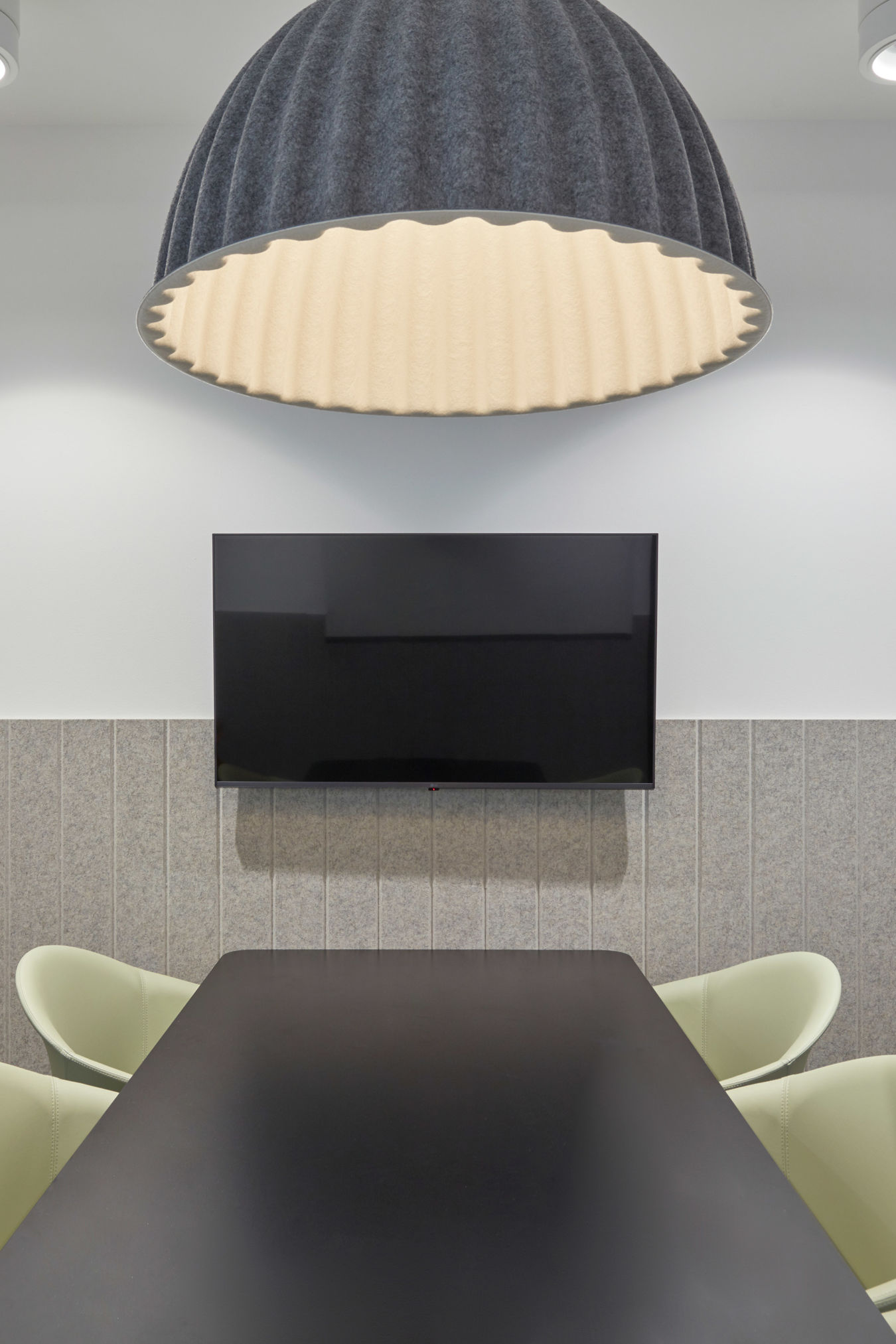 Modern conference room with a minimalist design featuring gray walls, a long black table, light green chairs, a large TV, and a ribbed dome light fixture