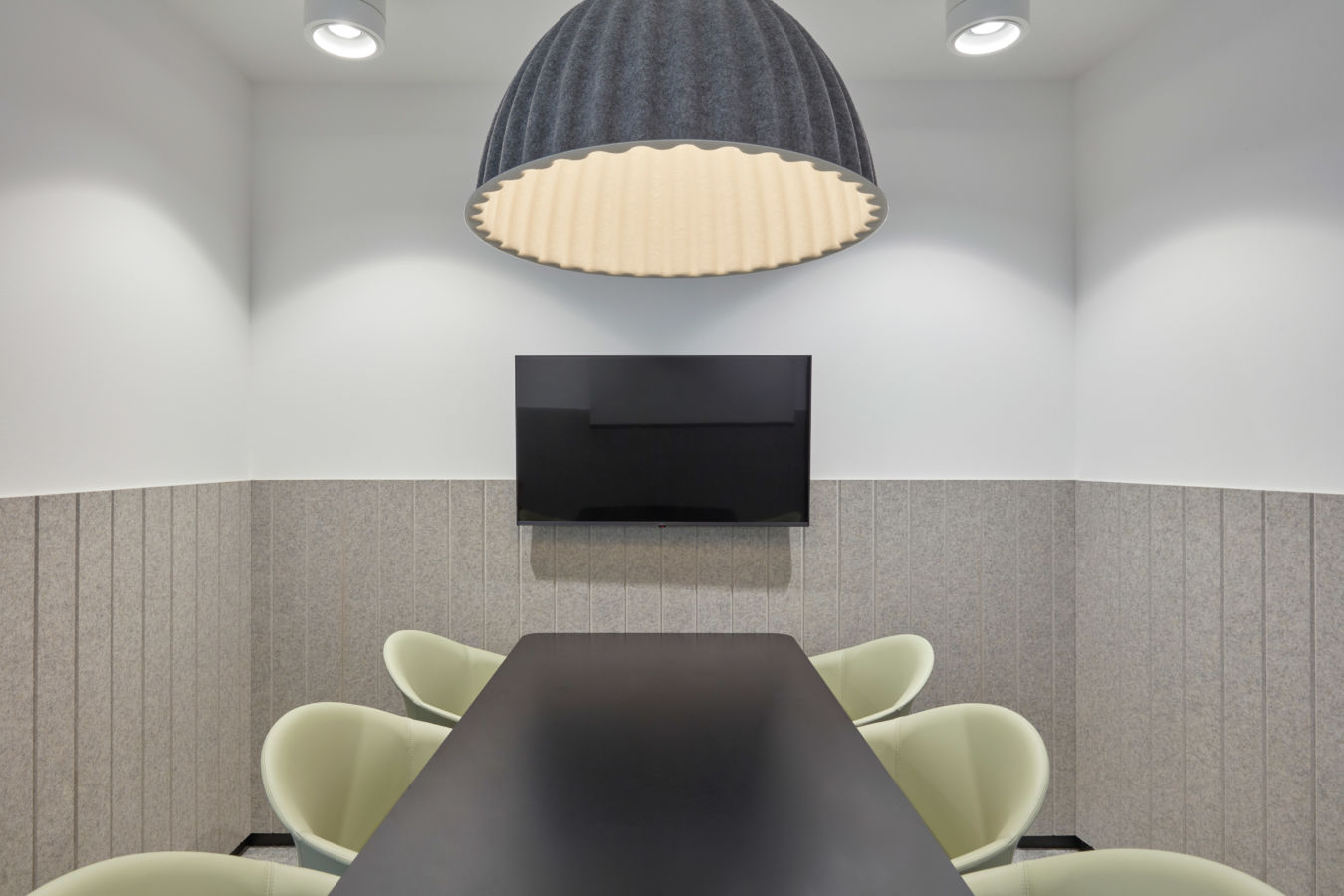 Modern conference room with a minimalist design featuring gray walls, a long black table, light green chairs, a large TV, and a ribbed dome light fixture