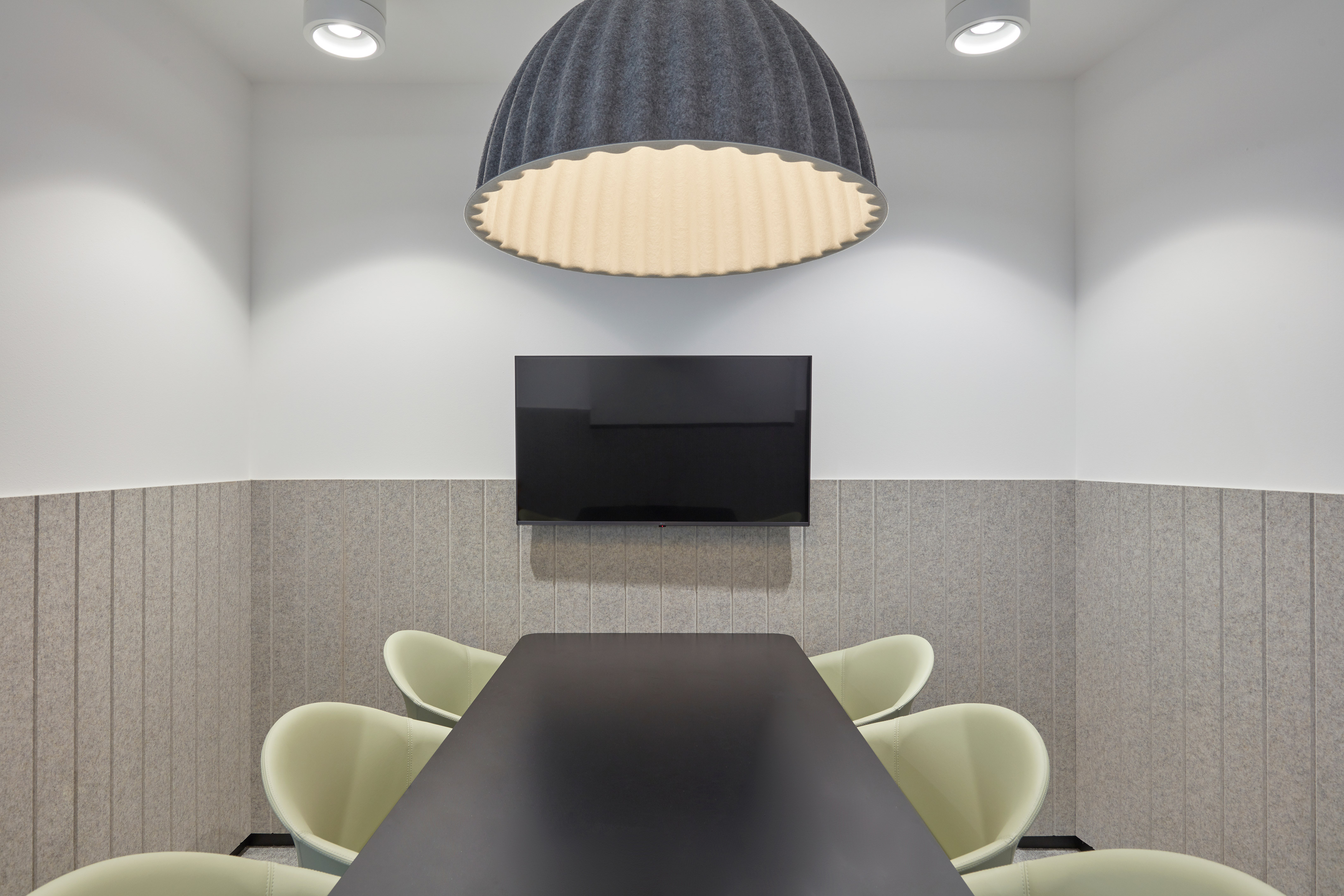 Modern conference room with a minimalist design featuring gray walls, a long black table, light green chairs, a large TV, and a ribbed dome light fixture