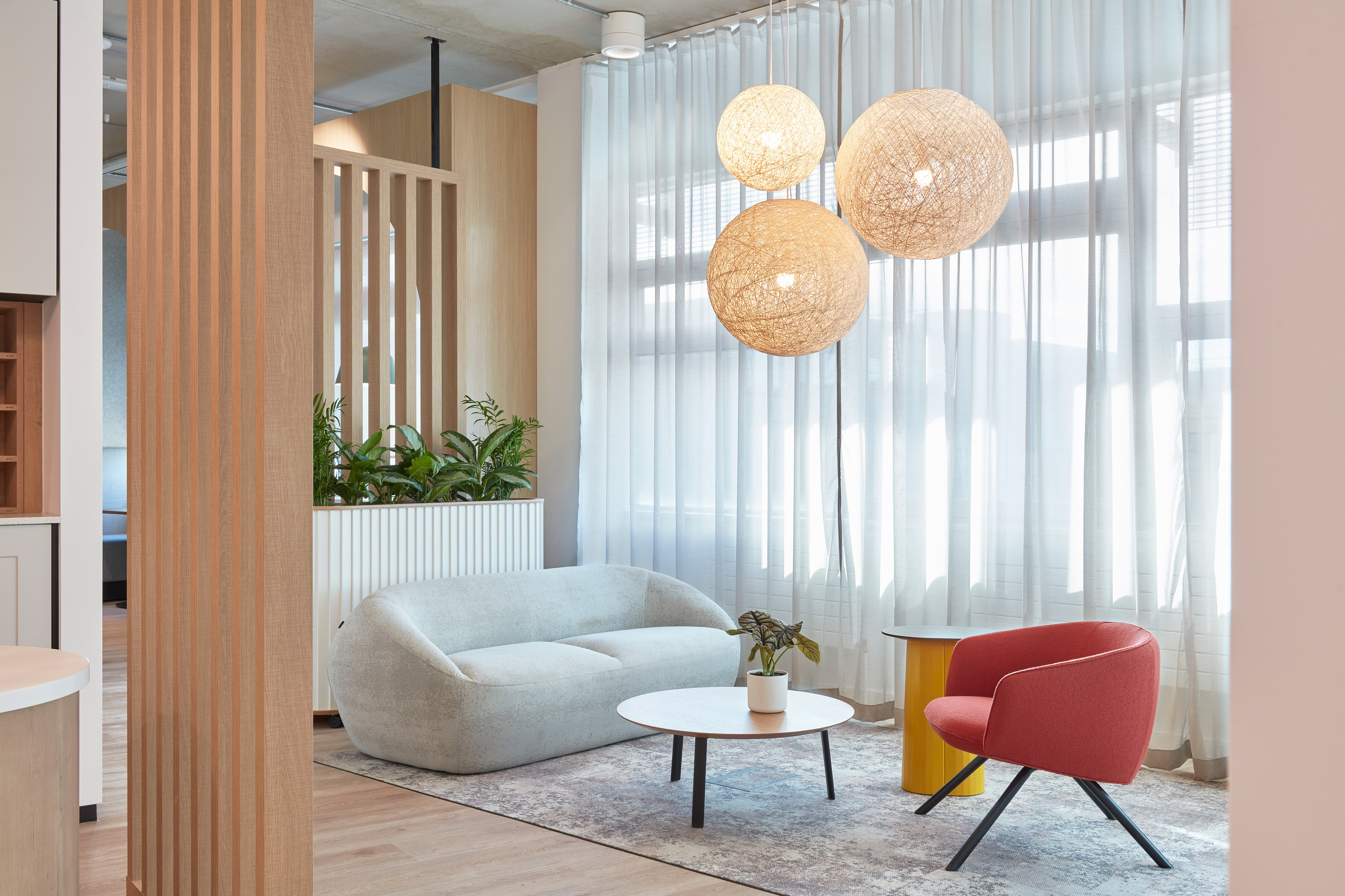 Modern living room with a light gray sofa and red armchair around a circular white table. Woven pendant lights hang above, soft natural light filters through sheer curtains