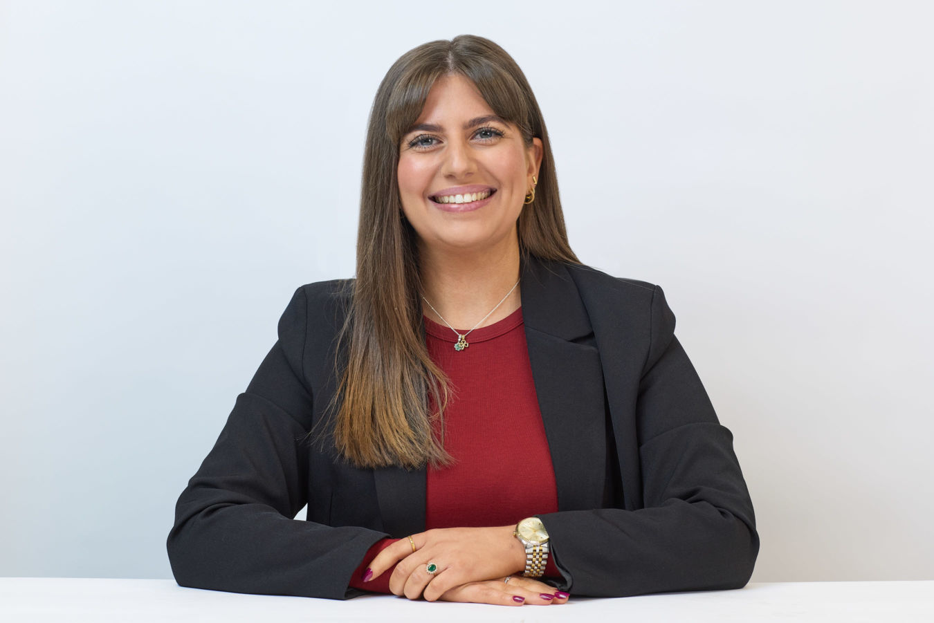 A person in a black blazer smiles while sitting at a white table, wearing a red top, gold watch, and necklace, set against a plain white background.