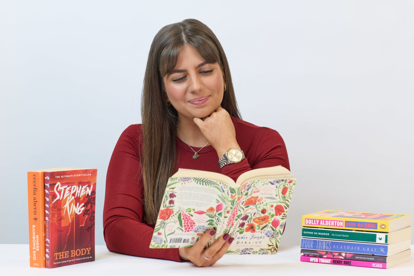 A woman sits at a table, reading a floral-covered book. Nearby, books by Stephen King and Dolly Alderton are stacked, creating a cozy reading ambiance.
