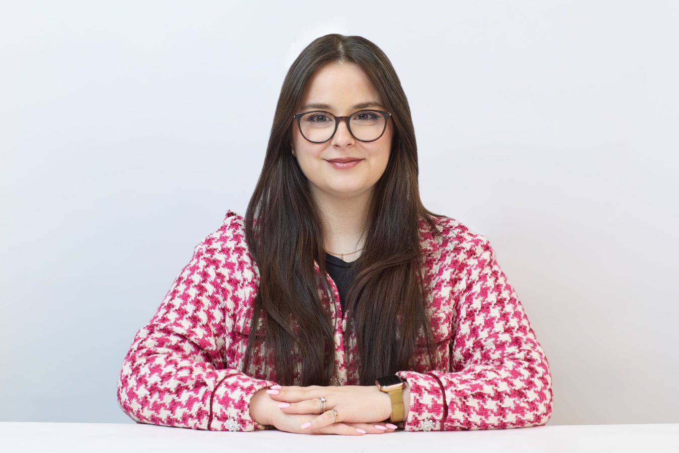 A person with long hair and glasses is sitting, resting hands on a surface, wearing a red patterned jacket. The background is plain white, emphasizing the individual.