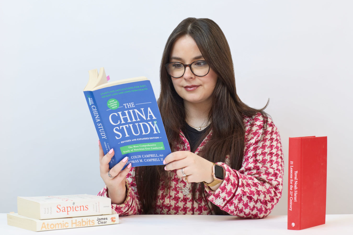A person in glasses reads "The China Study" book while sitting at a table; nearby are "Sapiens," "Atomic Habits," and "21 Lessons for the 21st Century" books.
