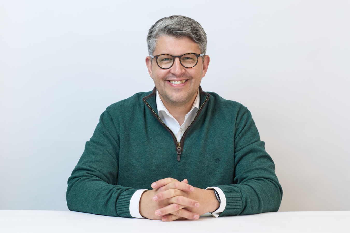 A person wearing glasses and a green pullover smiles while sitting at a white table against a plain background.