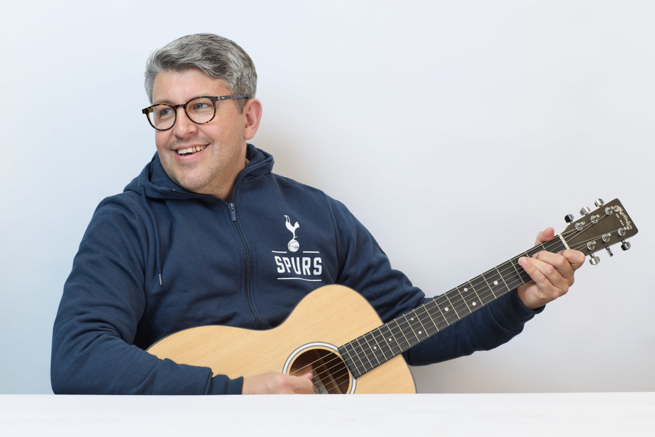 Man wearing glasses and a blue "Spurs" hoodie plays an acoustic guitar while smiling, seated at a white table against a plain background.