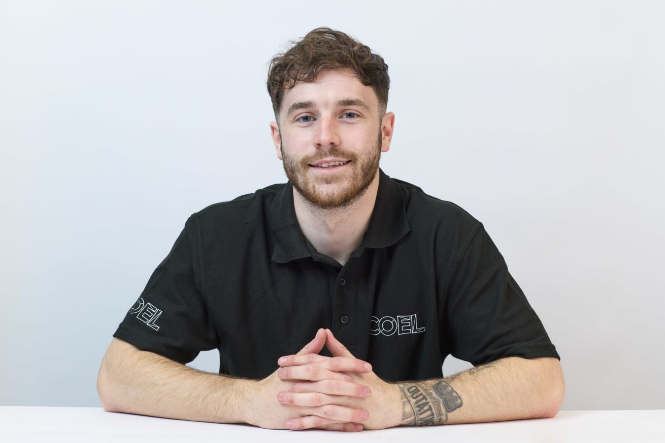 Young man sits at a white table, smiling, with hands clasped. He wears a black shirt embroidered with "COEL." The background is plain white. A tattoo is visible on his forearm.