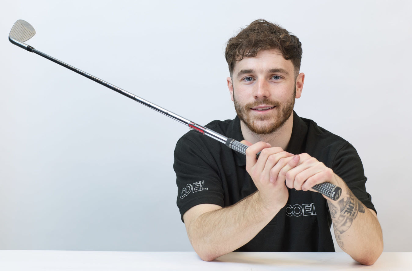 Man in a black shirt holding a golf club with both hands, smiling, in a bright indoor setting. Tattoo visible on his forearm. Shirt logo reads “COEL.”