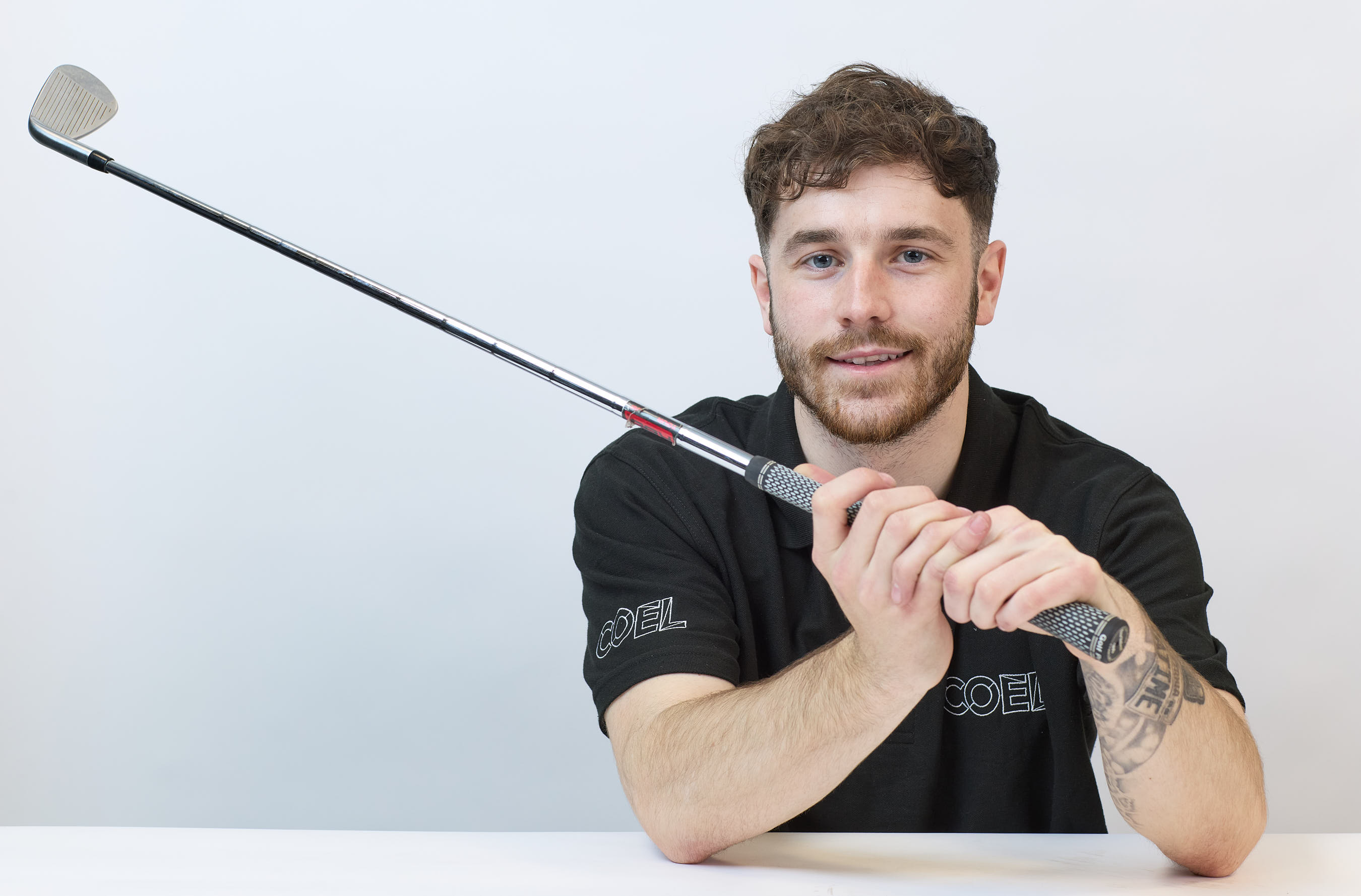 Man in a black shirt holding a golf club with both hands, smiling, in a bright indoor setting. Tattoo visible on his forearm. Shirt logo reads “COEL.”