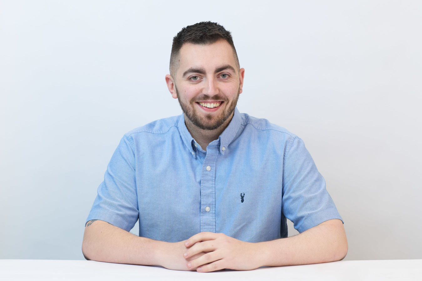 Smiling man in a light blue button-up shirt sits at a white table against a plain white background. His hands are clasped in front of him.