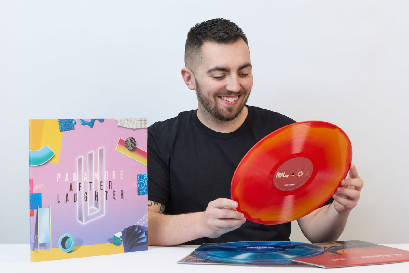 A person smiles while holding a red vinyl record. Beside them, a Paramore "After Laughter" album cover rests on a table. A blue vinyl record lies beneath.