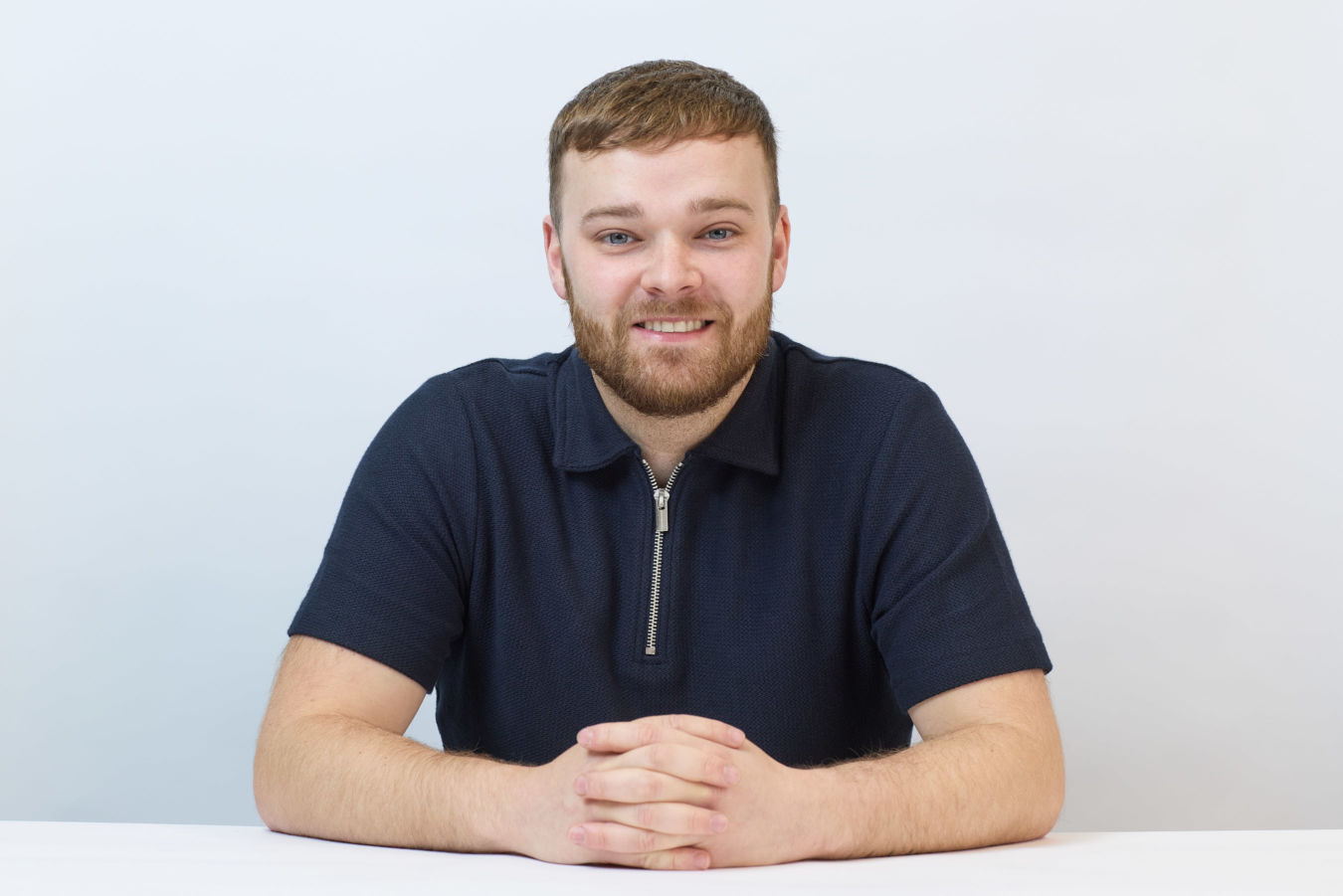 A person sits smiling with hands clasped on a white table, wearing a dark zip-up shirt against a plain white background.