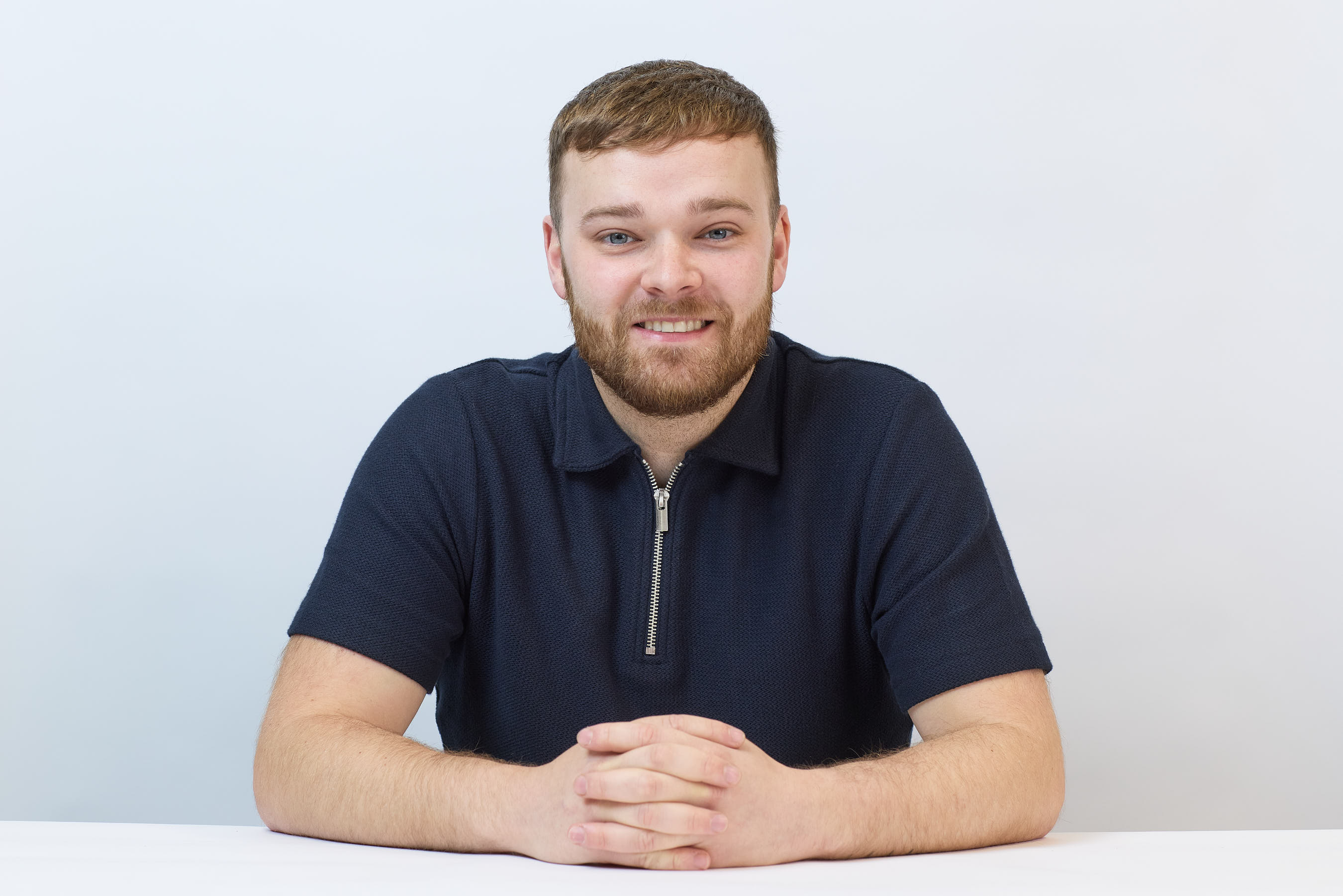 A person sits smiling with hands clasped on a white table, wearing a dark zip-up shirt against a plain white background.
