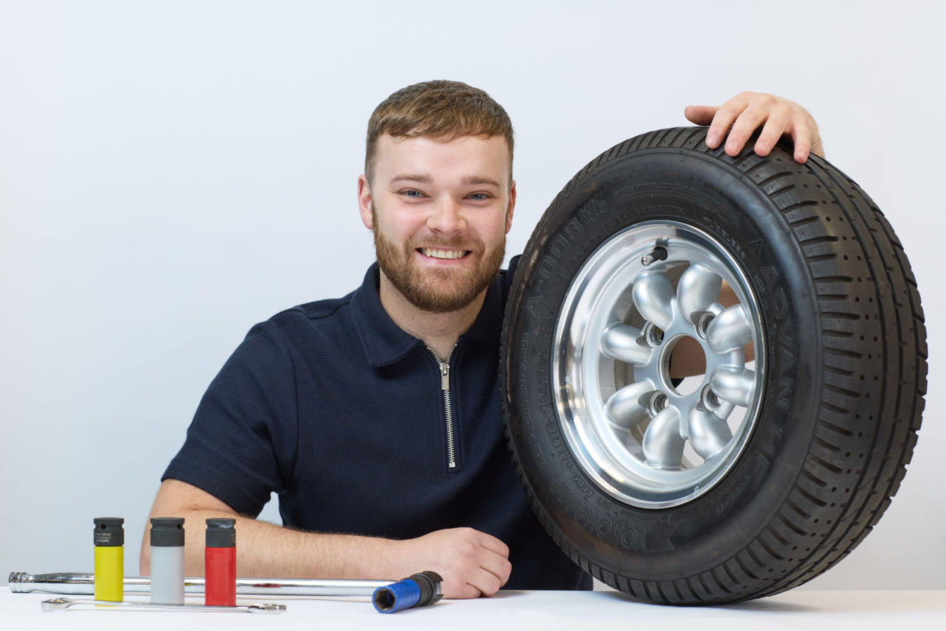 A smiling person holds a large car tire, labeled "Yokohama," on a table with three colorful cylinder tools and metallic rods against a plain white background.