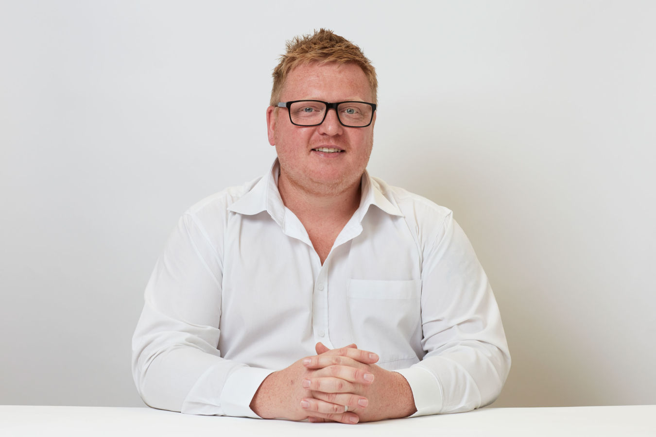 A person wearing glasses and a white shirt sits at a table with hands clasped, against a plain white background.