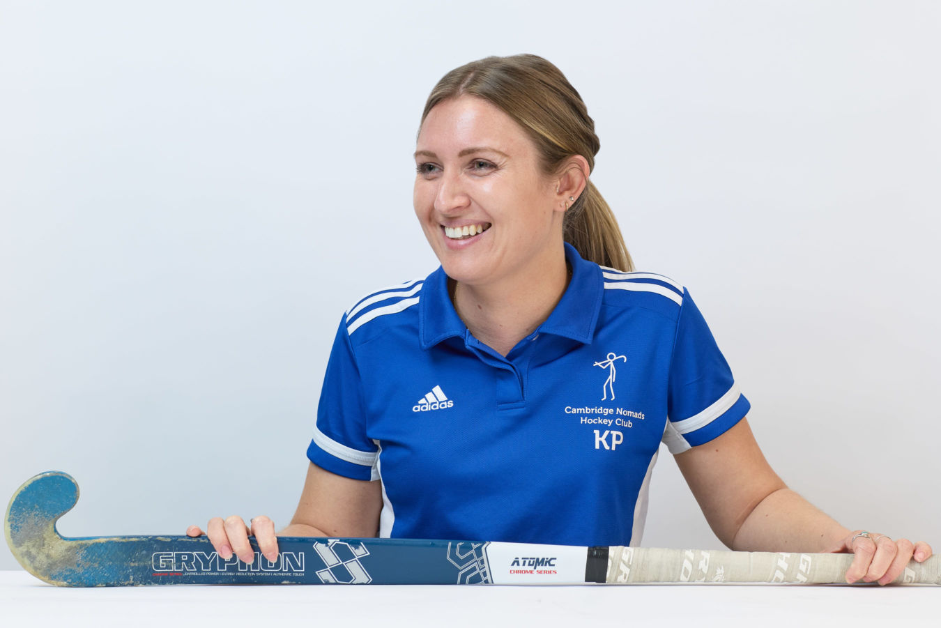 A person in a blue "Cambridge Nomads Hockey Club" shirt holds a Gryphon field hockey stick, smiling in a well-lit, neutral setting. The shirt displays Adidas and KP logos.