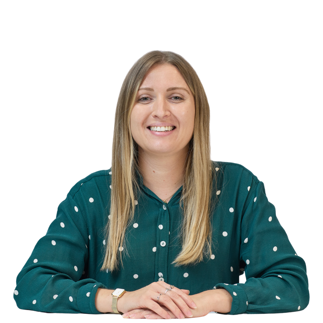 Smiling woman sits at a table, wearing a green polka dot blouse, with hands resting in front; white background.