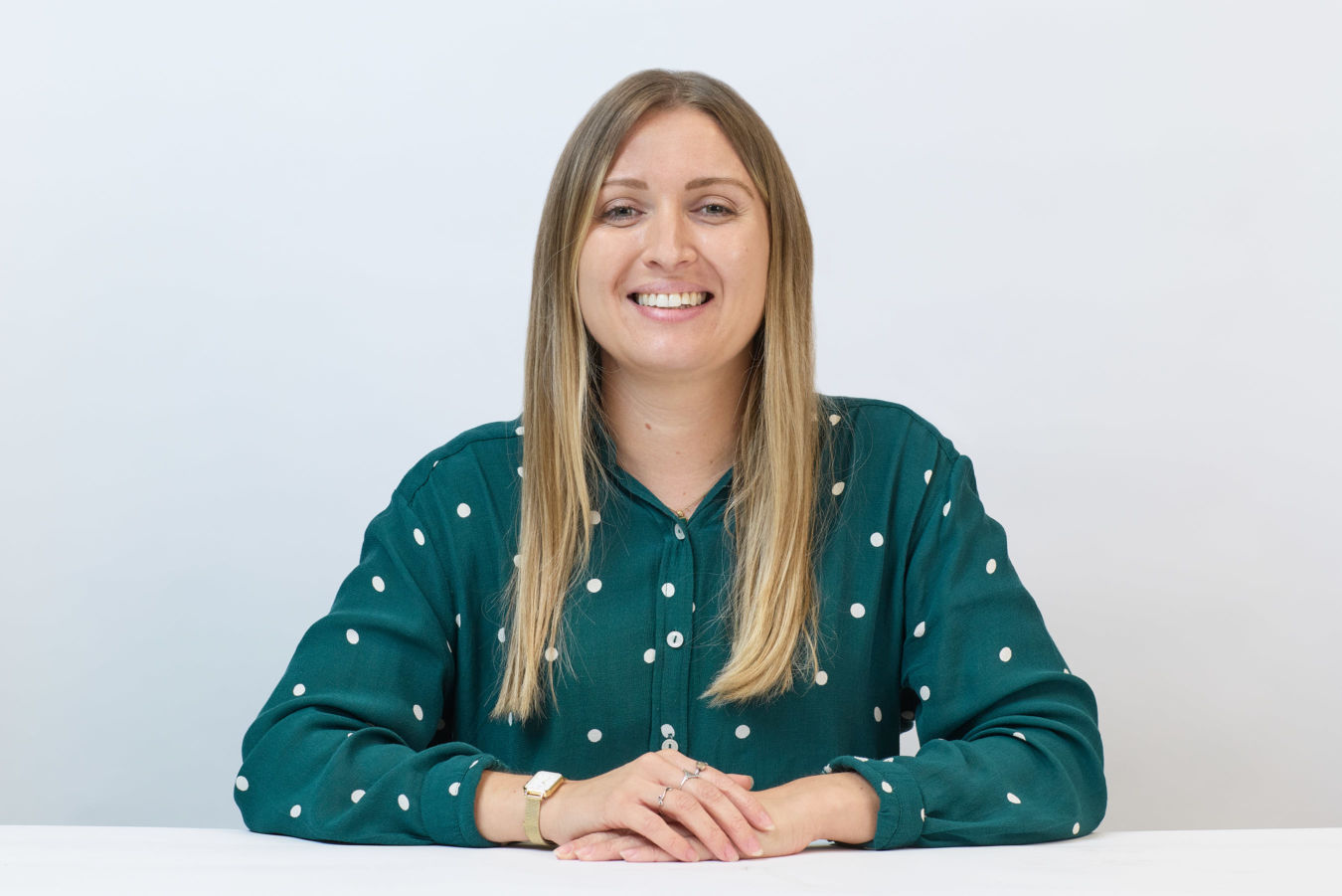 Smiling woman sits at a table, wearing a green polka dot shirt, against a plain white background, hands resting in front.