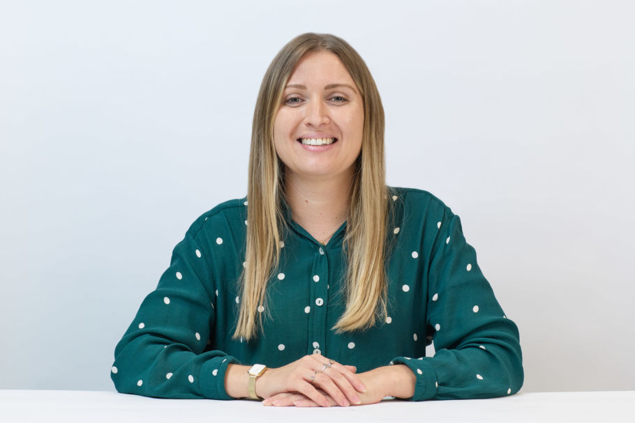 Smiling woman sits at a table, wearing a green polka dot shirt, against a plain white background, hands resting in front.