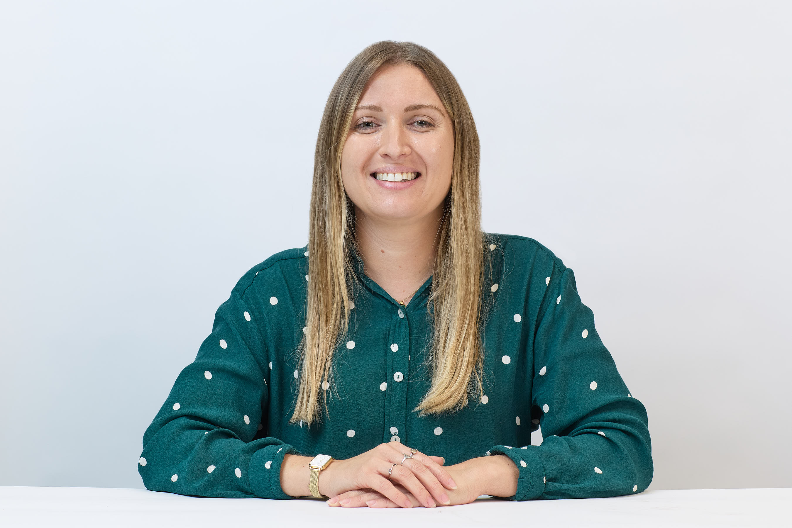 Smiling woman sits at a table, wearing a green polka dot shirt, against a plain white background, hands resting in front.