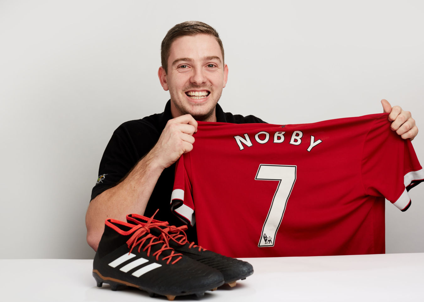 A person holds a red jersey with "NOBBY 7" printed on it, smiling, beside black soccer cleats on a white table, against a plain white background.