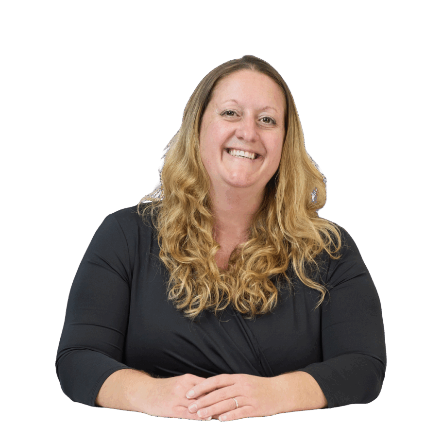 A smiling person with long, wavy hair sits at a table, wearing a black top, hands gently clasped, against a plain white background.