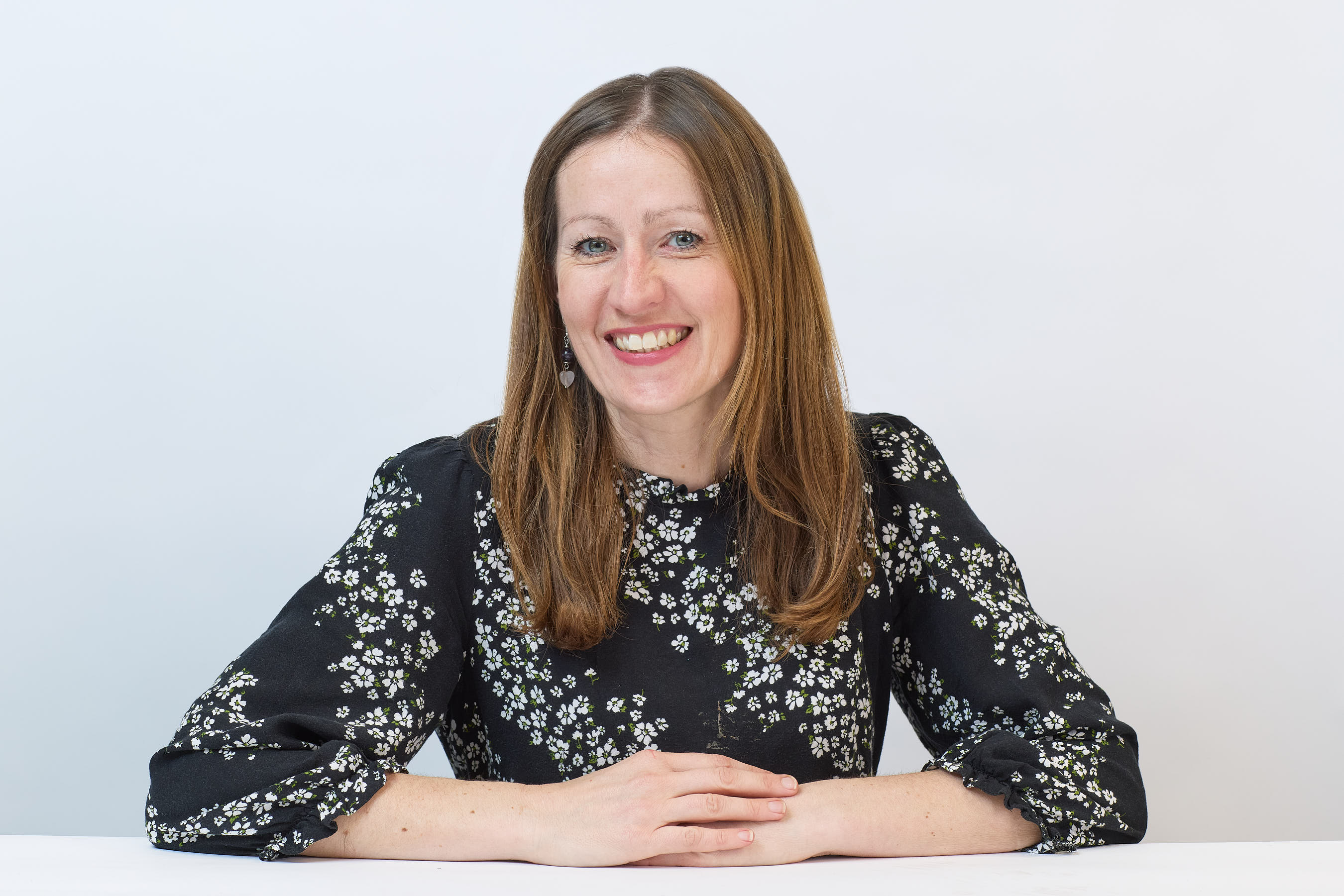 Smiling woman sits with hands folded on a table, wearing a black and white floral patterned blouse, set against a plain light background.