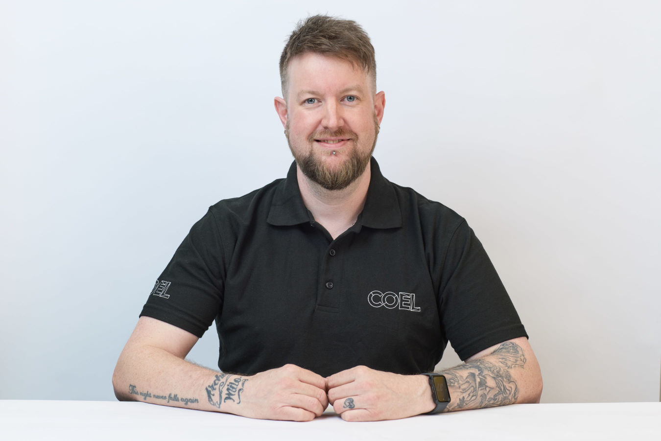 A person sits at a white table, wearing a black polo shirt with "COEL" on it, in front of a plain white background. Tattoos are visible on their arms.