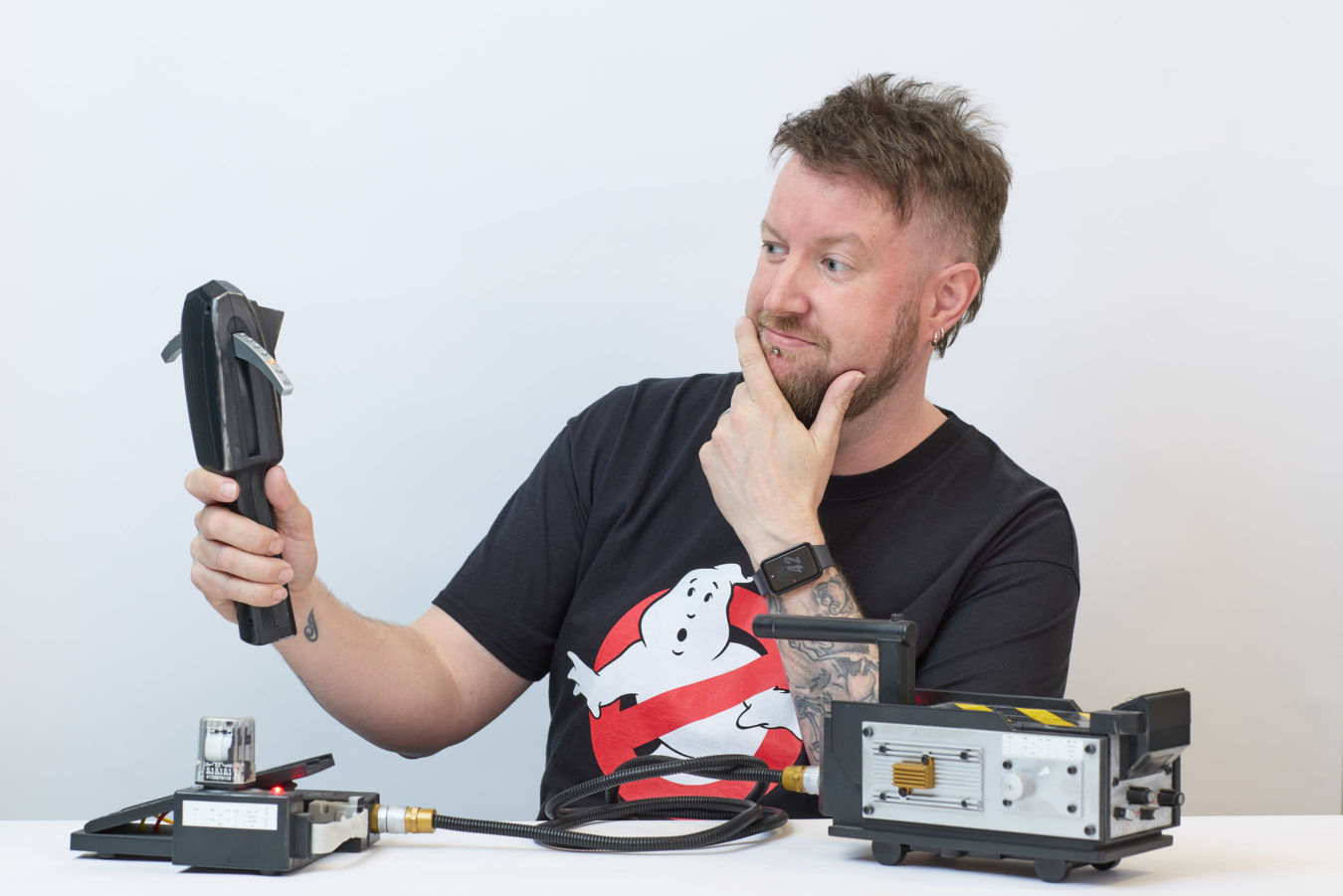 A person holds a black device inquisitively while seated at a table with electronic gadgets. They wear a Ghostbusters t-shirt and smartwatch, appearing thoughtful against a plain background.