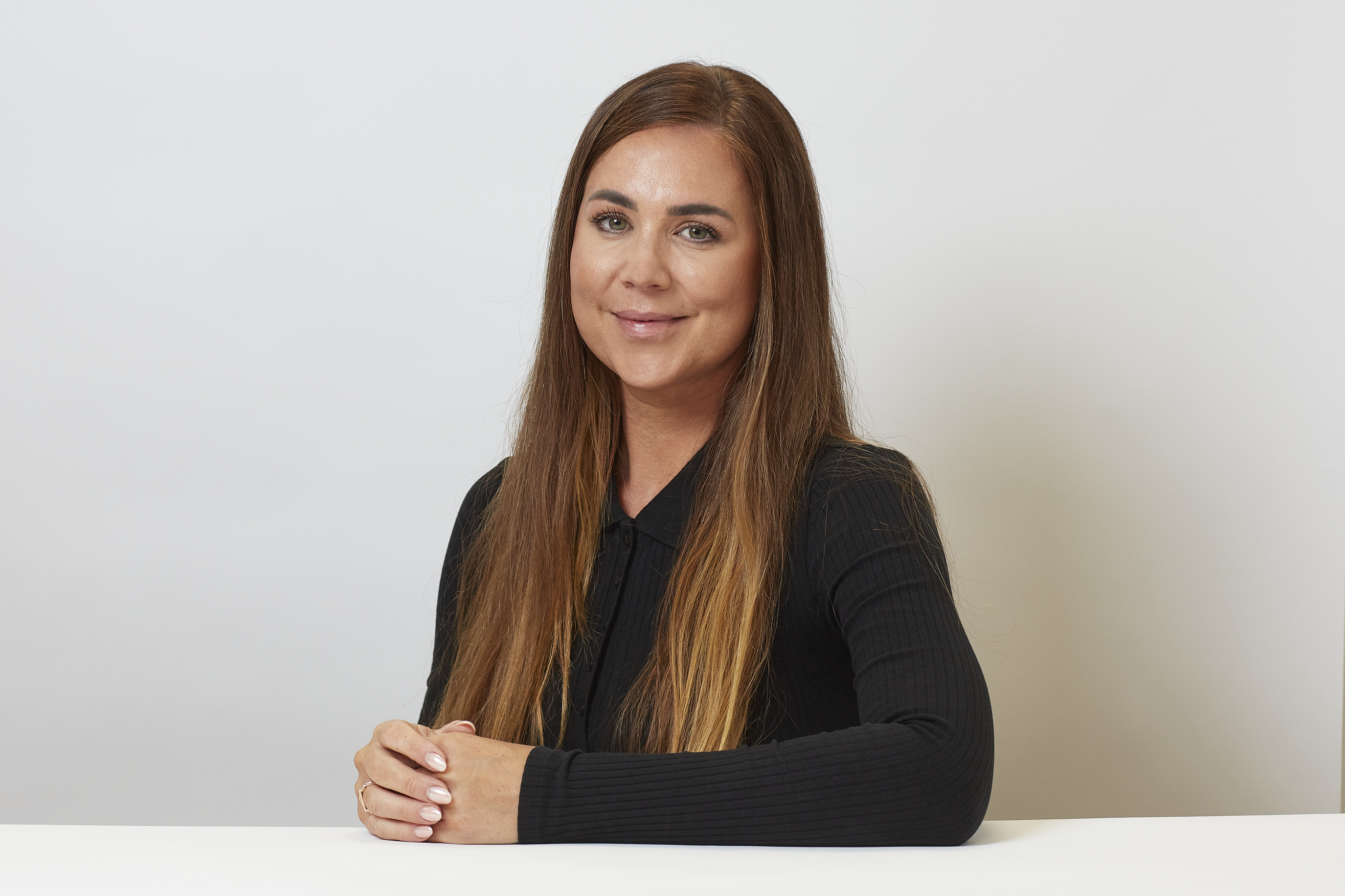 A woman with long brown hair sits at a table, hands clasped, against a plain white background. She wears a black long-sleeve top and smiles gently.