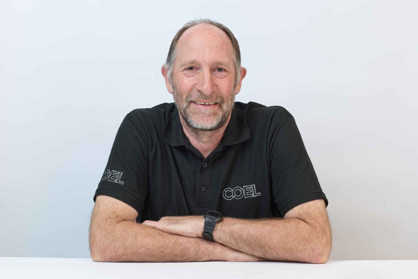 Man smiling with arms crossed, wearing a black polo shirt with "COEL" on the sleeve and chest, in front of a plain white background.