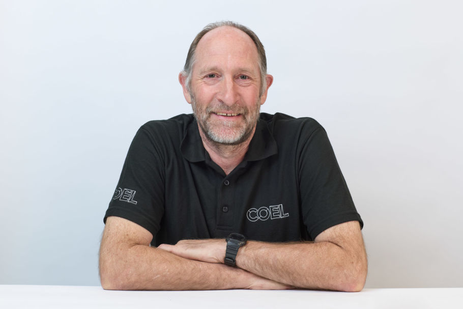 Man smiling with arms crossed, wearing a black polo shirt with "COEL" on the sleeve and chest, in front of a plain white background.