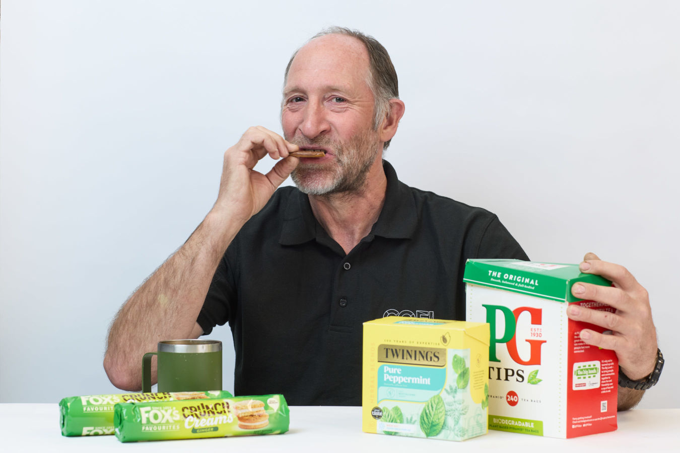 A man in a black shirt eats a biscuit, holding a PG Tips tea box, with Twinings Peppermint tea and Fox's Crunch Creams biscuits on a white table.